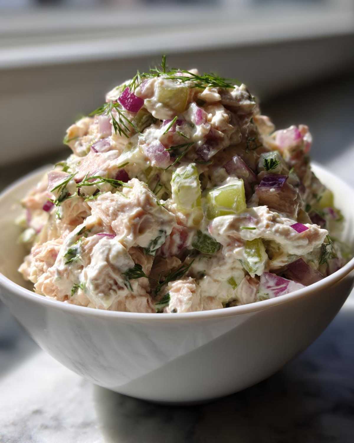 A mound of creamy, chunky healthy tuna salad mixed with red onion and celery, garnished with fresh dill, served in a white bowl.