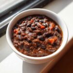 Close-up of a small white bowl filled with thick, rich black bean chili, highlighted by sunlight.