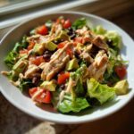 Close-up of a vibrant southwest salad featuring grilled chicken strips, avocado, black beans, tomatoes, and creamy dressing.