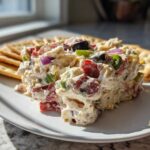 A scoop of creamy hoagie dip loaded with colorful vegetables and served with crackers on a white plate.