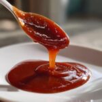 Close-up of thick, rich homemade bbq sauce being drizzled from a white spoon onto a white plate.