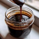 A spoonful of thick, dark chocolate syrup drips back into a glass jar of homemade chocolate syrup.