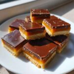 Close-up of several homemade twix bites stacked on a white plate, showing shortbread, caramel, and chocolate layers.