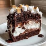 A decadent slice of hot chocolate poke cake layered with cream and topped with chocolate drizzle and toasted marshmallows.