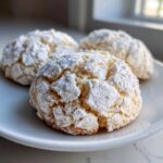Three freshly baked lemon crinkle cookies heavily dusted with powdered sugar resting on a white plate.