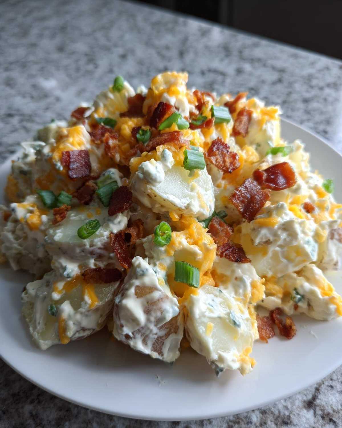 A close-up of loaded baked potato salad featuring chunks of potato coated in creamy dressing, topped with cheddar cheese, bacon bits, and green onions.