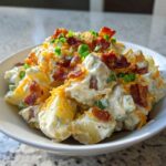 A close-up of creamy baked potato salad topped with shredded cheddar, crispy bacon bits, and green onions.