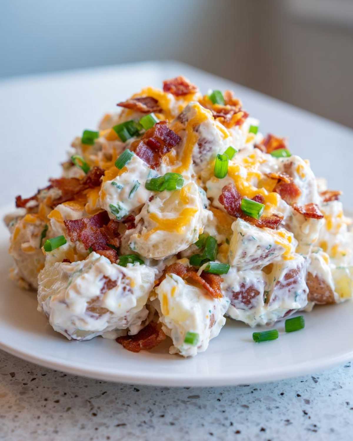 A mound of creamy baked potato salad topped with shredded cheddar cheese, crispy bacon bits, and fresh green onions.
