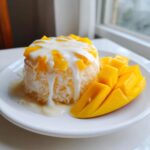 A serving of mango sticky rice topped with coconut cream and fresh mango slices on a white plate.