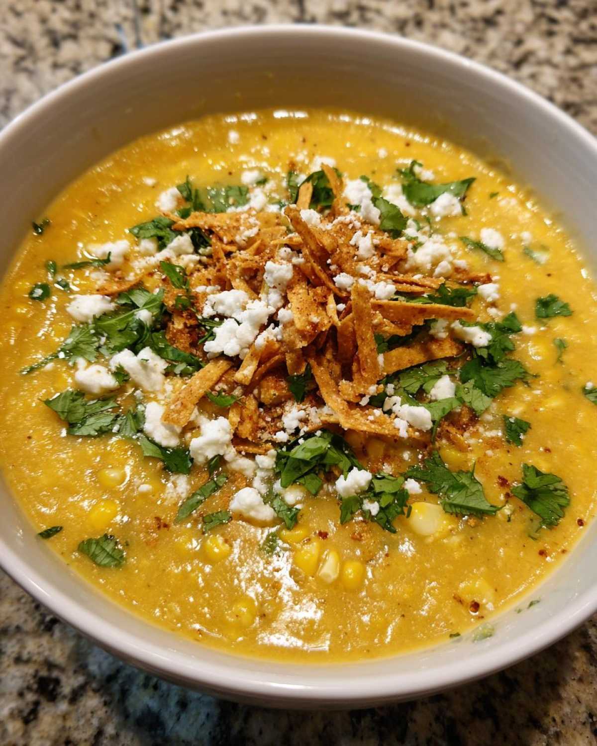 A close-up view of a bowl of creamy, yellow Mexican street corn soup topped with cotija cheese, cilantro, and tortilla strips.