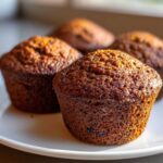 Four freshly baked, dark brown bran muffins sitting closely together on a white plate, highlighted by natural light.