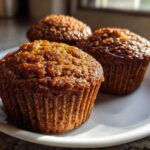 Close-up of three golden-brown, moist bran muffins sitting on a white plate near a window.
