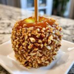 A close-up of a glossy caramel apples dipped in chopped nuts, served on a white square plate.