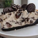 A thick slice of creamy Oreo cheesecake with a dark chocolate crust, topped with crushed and whole Oreo cookies.