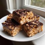 Four squares of peanut butter rice krispie treats topped with melted chocolate drizzle stacked on a white plate.
