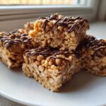 A stack of homemade peanut butter rice krispie treats drizzled with melted chocolate, sitting on a white plate.