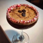 A close-up of a peppermint espresso martini garnished with a crushed candy cane rim and three coffee beans.