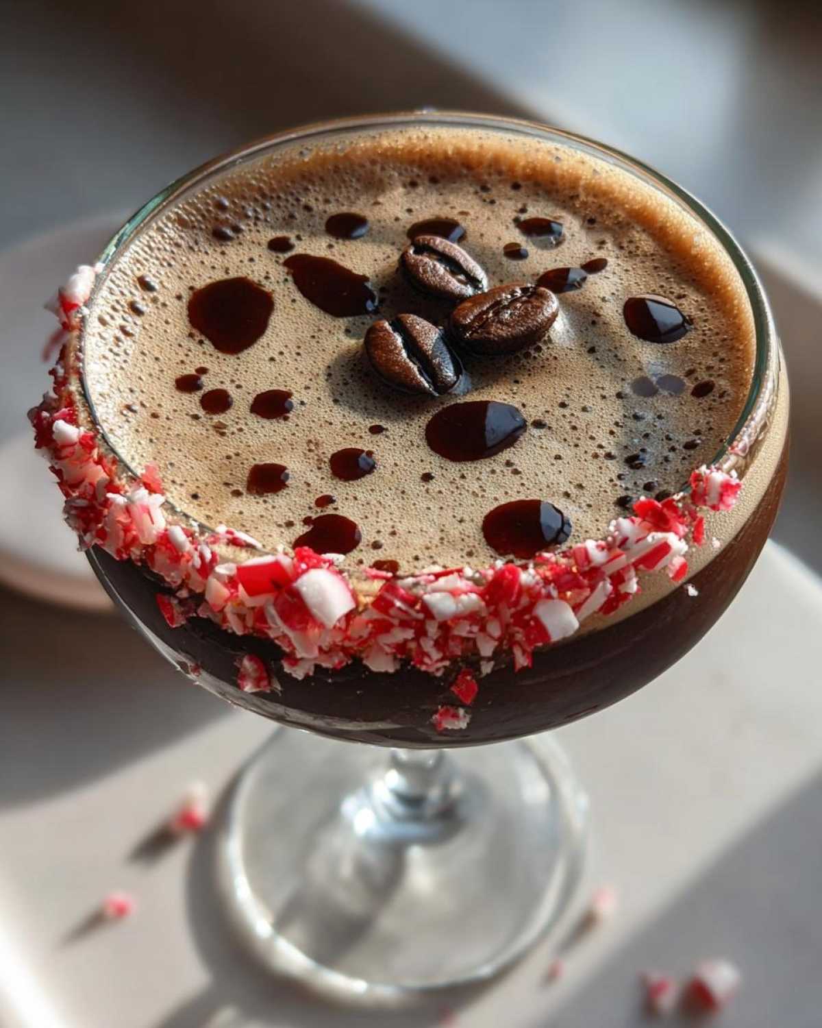 Close-up of a peppermint espresso martini garnished with a crushed candy cane rim, coffee beans, and chocolate syrup.