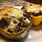 Two baked philly cheesesteak stuffed peppers, one green and one yellow, topped with melted, bubbly cheese.