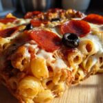 A close-up, cross-section view of a slice of baked pizza pasta casserole topped with melted cheese, pepperoni, and a black olive.