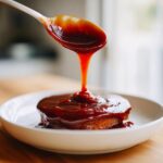 A spoonful of thick, glossy homemade bbq sauce is being poured over a small, round piece of food on a white plate.