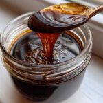 A spoonful of rich, dark homemade chocolate syrup drips back into a glass jar, showing its thick consistency.