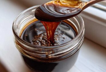 A spoonful of rich, dark homemade chocolate syrup drips back into a glass jar, showing its thick consistency.