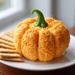A festive orange pumpkin cheese ball shaped like a small pumpkin, topped with a green pepper stem, served with crackers.