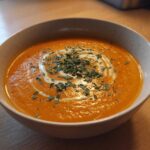 A close-up of a bowl of vibrant orange roasted butternut squash soup, garnished with a swirl of cream and fresh herbs.