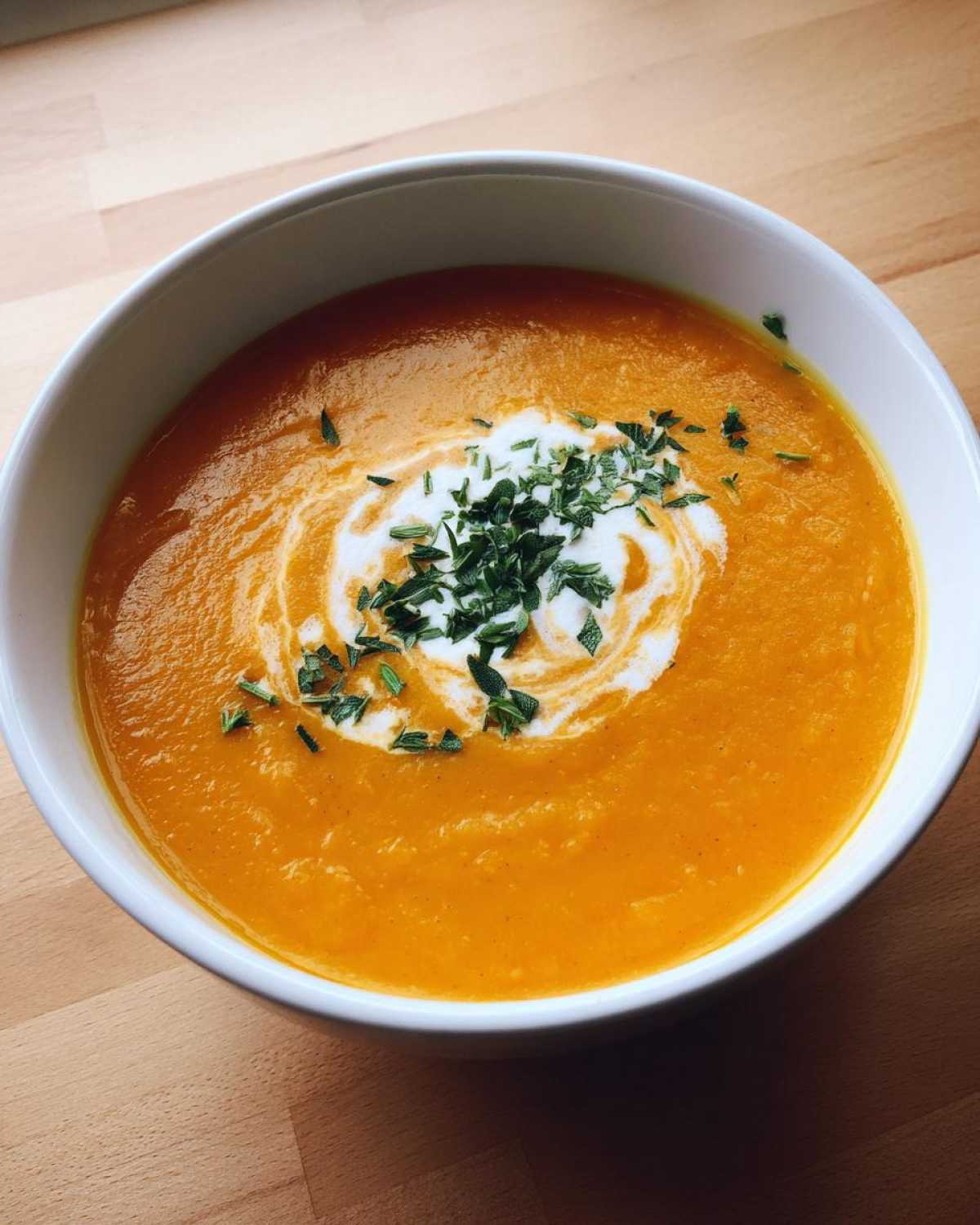 A close-up view of a white bowl filled with vibrant orange roasted butternut squash soup, garnished with a swirl of cream and fresh herbs.