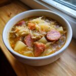 Close-up of a white bowl filled with steaming sauerkraut soup featuring chunks of sausage and potatoes.