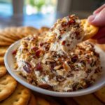 A hand scoops a generous portion of creamy Neiman Marcus dip topped with bacon and almonds onto a round cracker.