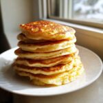 A tall stack of six perfectly cooked, fluffy pancakes with golden brown tops sitting on a white plate.