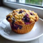 Close-up of a freshly baked blueberry oatmeal muffin topped with coarse sugar, sitting on a white plate.