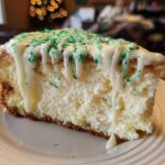A close-up of a slice of creamy christmas tree cheesecake topped with white icing drizzle and green sprinkles.
