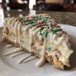 A close-up of a slice of christmas tree cheesecake topped with white chocolate drizzle and festive red and green sprinkles.