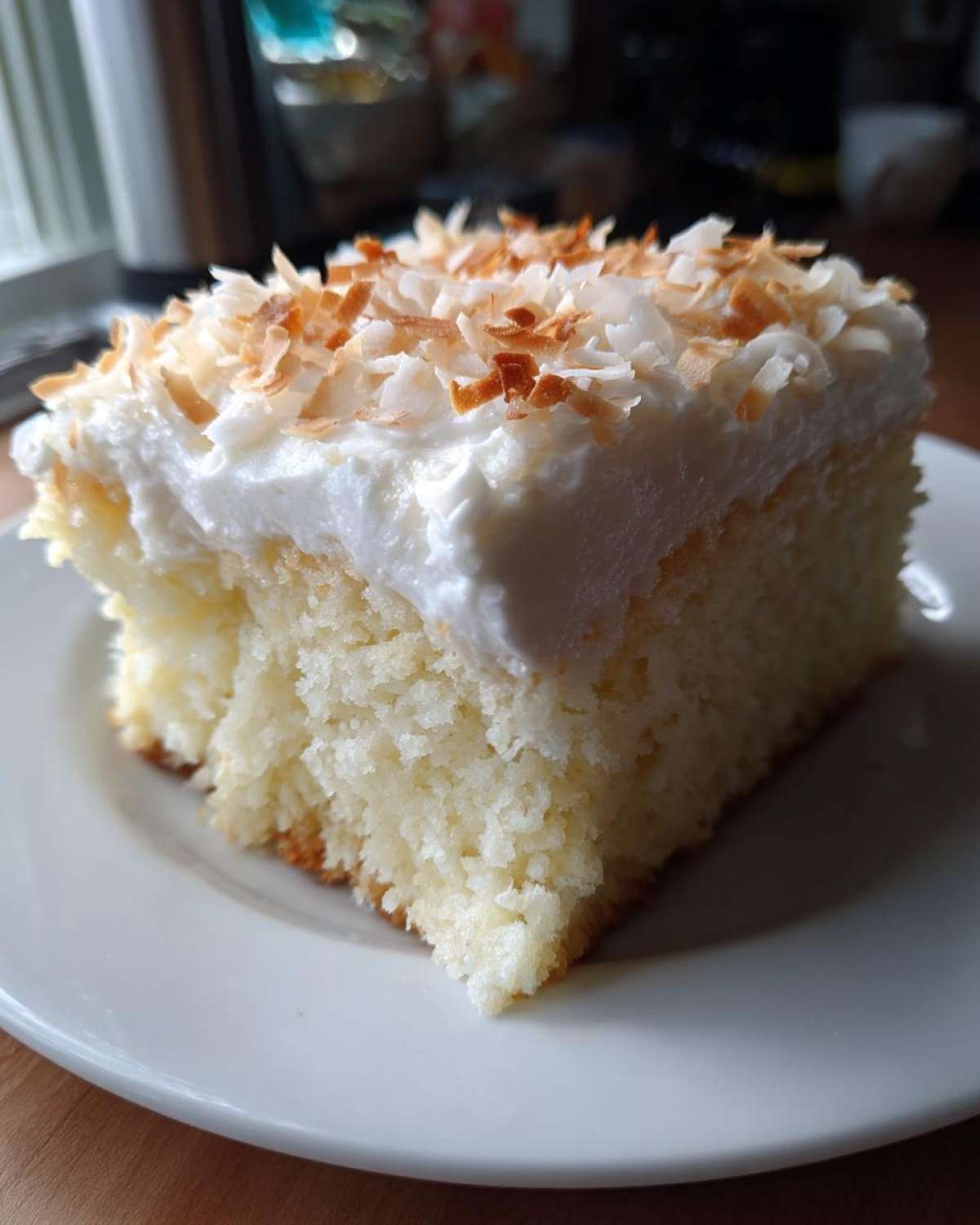 A close-up of a moist slice of coconut poke cake topped with white frosting and toasted coconut flakes.