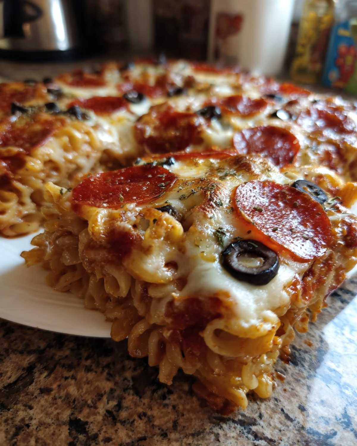 Close-up of a slice of baked pizza pasta casserole topped with melted cheese, pepperoni, and olives.