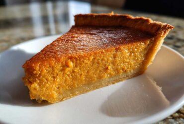 A close-up, sunlit slice of rich, orange sweet potato pie resting on a white dessert plate.