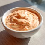 A white bowl filled with creamy, orange-tinted smash burger sauce speckled with red spices, sitting on a wooden surface.