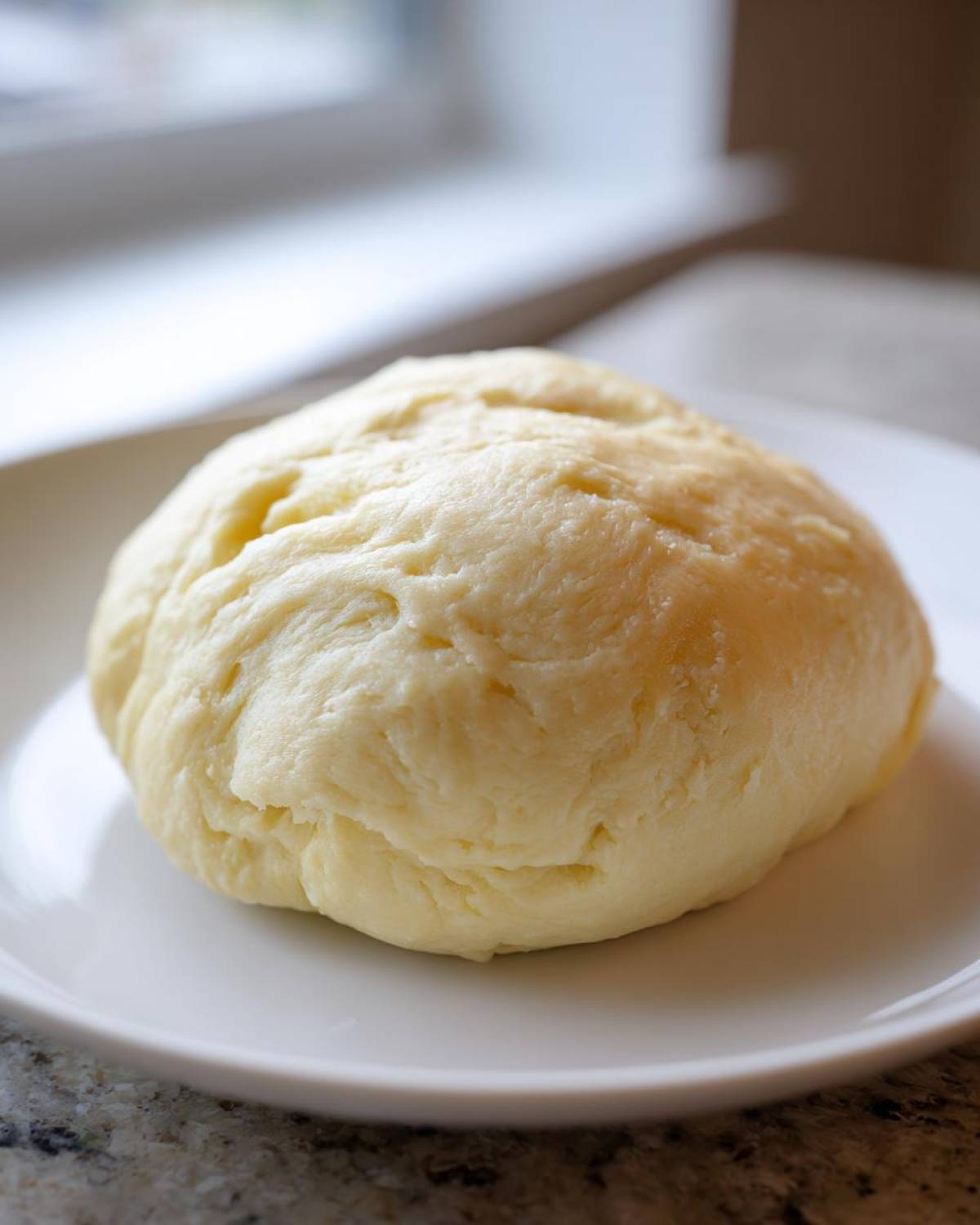 A close-up of a freshly made, smooth, pale yellow ball of fufu resting on a white plate.