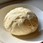 A perfectly smooth, pale yellow ball of freshly made fufu resting on a white plate.