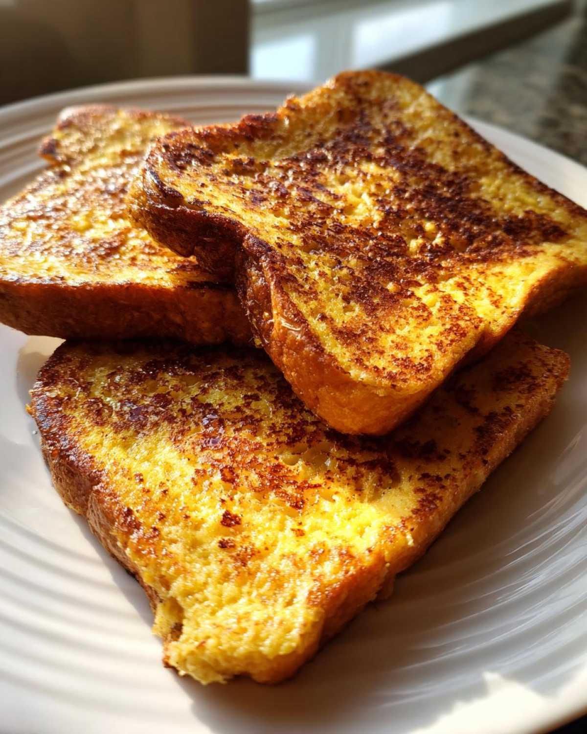 A close-up of three golden-brown slices of sourdough french toast stacked on a white plate.