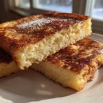 Close-up of thick, golden-brown sourdough french toast slices stacked on a white plate, dusted with powdered sugar.