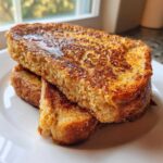 Close-up of thick, golden-brown sourdough french toast stacked on a white plate, drizzled with syrup.