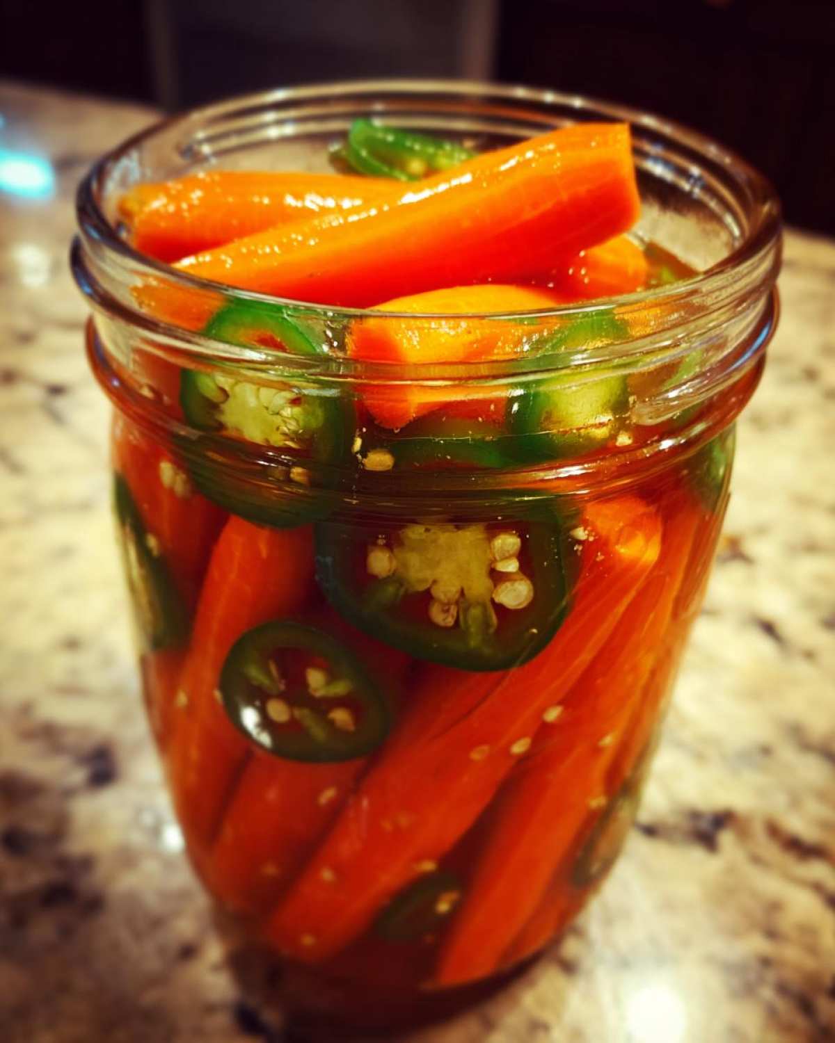 Close-up of bright orange pickled carrots and sliced green jalapeños packed tightly in a glass jar.
