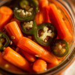 Close-up of bright orange pickled carrots submerged in liquid with slices of green jalapeño peppers.