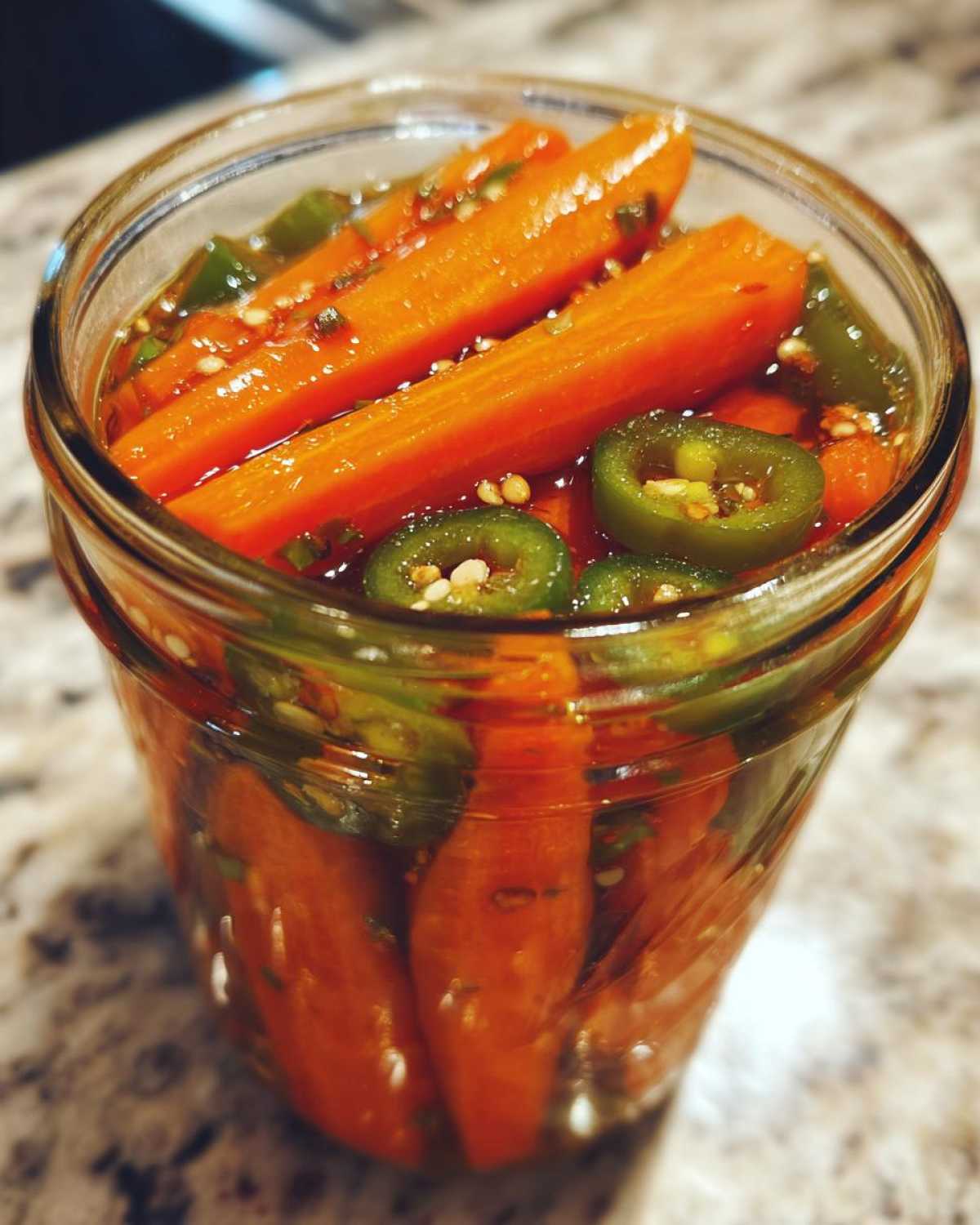 Close-up of bright orange pickled carrots and sliced jalapeños submerged in brine inside a glass jar.