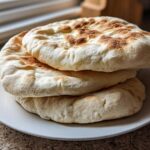 A stack of three fluffy, freshly baked pita bread rounds with golden-brown spots resting on a white plate.