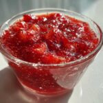 Close-up of vibrant red strawberry freezer jam with visible seeds, served in a small clear glass bowl.